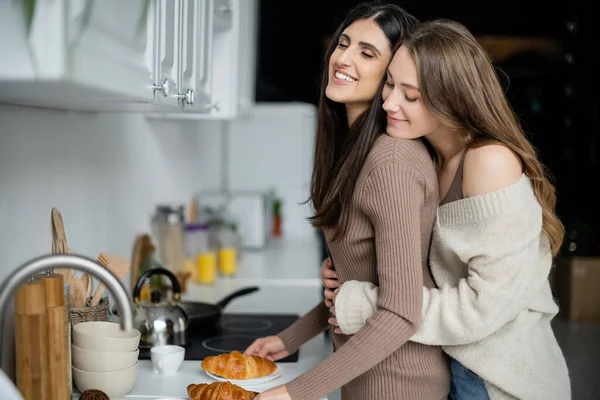 Позитивная лесбиянка в уютном свитере обнимает подружку возле круассанов на кухне — стоковое фото