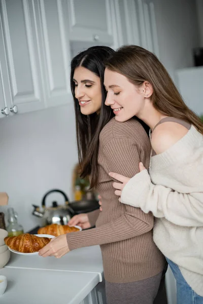 Smiling woman in sweater hugging girlfriend with tasty croissants in kitchen — Stock Photo