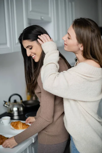 Vista lateral da mulher em suéter tocando o cabelo da namorada com croissants na cozinha — Fotografia de Stock