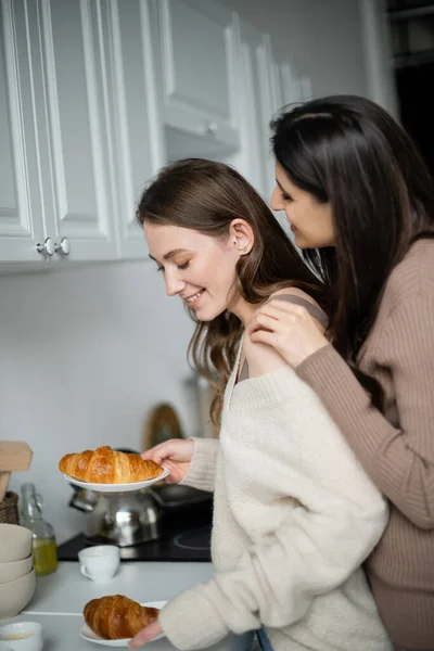 Young woman hugging partner in cozy sweater holding croissants in kitchen — Stock Photo