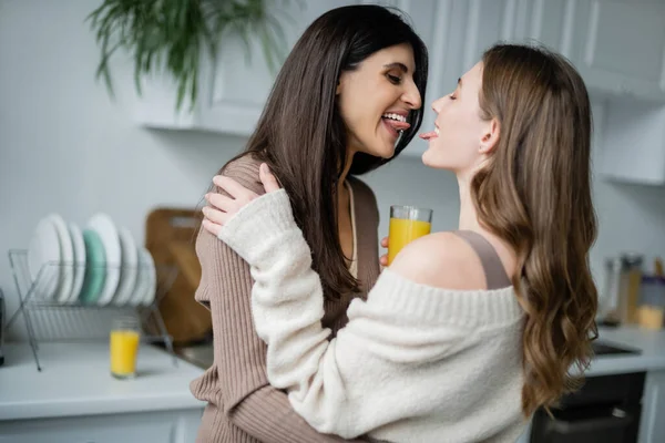 Lesbian women with orange juice sticking out tongues in kitchen — Stock Photo