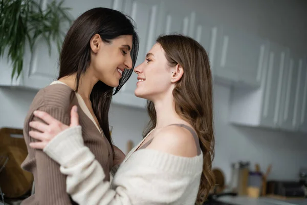 Vista lateral de mujeres lesbianas alegres abrazándose en cocina borrosa - foto de stock