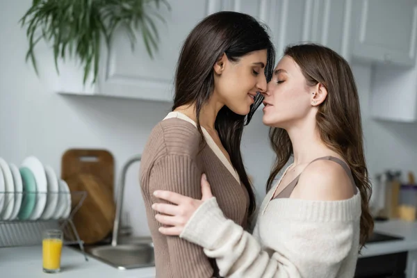 Side view of woman in sweater kissing brunette girlfriend in kitchen — Stock Photo