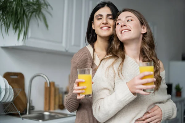 Mujer alegre sosteniendo jugo de naranja y abrazando novia en la cocina - foto de stock