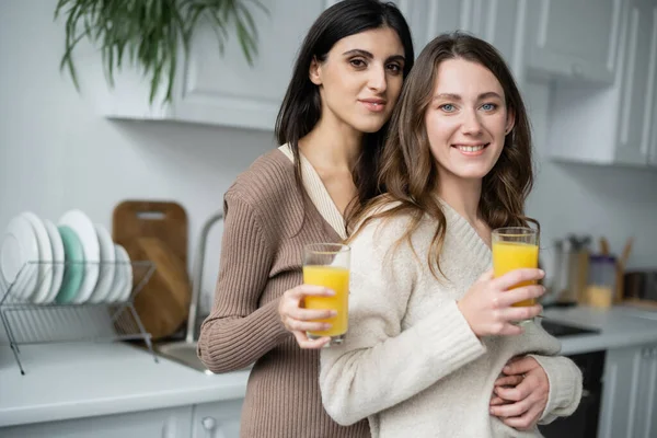 Улыбающаяся лесбийская пара держит стаканы апельсинового сока на кухне — стоковое фото