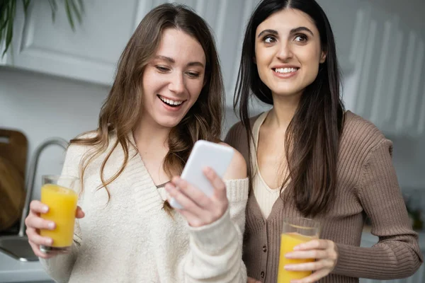 Mujer sonriente usando smartphone y sosteniendo jugo de naranja cerca de su novia en la cocina - foto de stock
