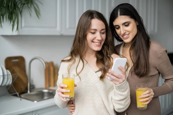 Same sex couple holding orange juice and using smartphone in kitchen — Stock Photo