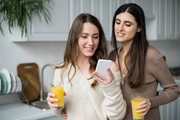 Young woman holding orange juice and using smartphone near girlfriend in kitchen at home — Stock Photo