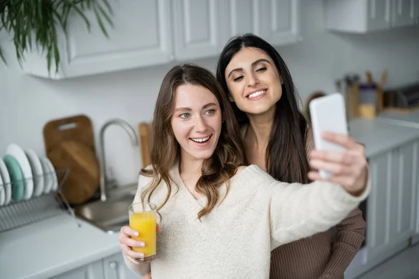 Усміхнена лесбійська пара з апельсиновим соком приймає селфі на смартфон на кухні — стокове фото