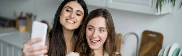 Positive same sex couple taking selfie on smartphone in blurred kitchen, banner — Stock Photo