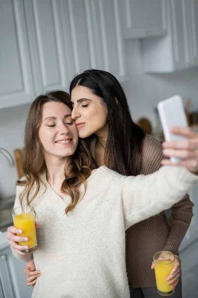 Mujer lesbiana sosteniendo jugo de naranja y besando a su novia mientras toma selfie en la cocina - foto de stock