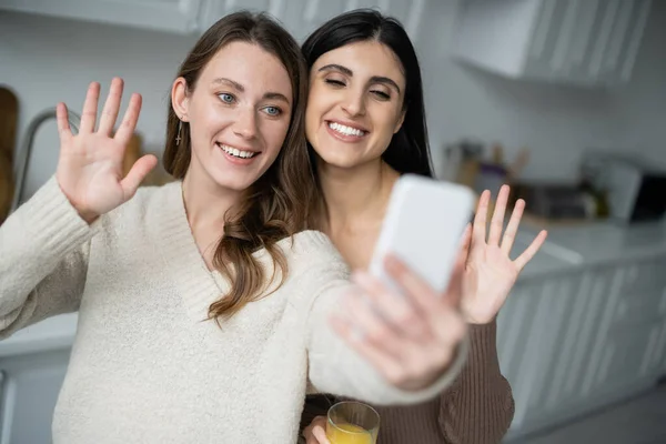 Улыбающаяся лесбийская пара машет руками во время видеозвонка на смартфон на кухне — стоковое фото