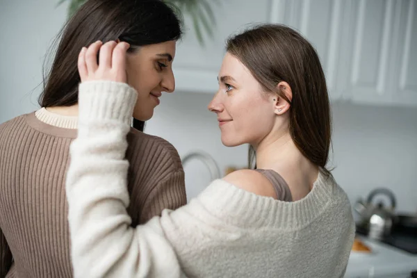 Side view of smiling woman in sweater touching hair of girlfriend in kitchen — Stock Photo