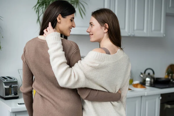 Vista lateral de jovens lésbicas cupê abraçando e olhando uns para os outros na cozinha — Fotografia de Stock