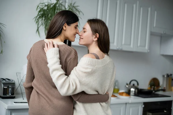 Вид сбоку на улыбающуюся лесбийскую пару, обнимающуюся и стоящую носом к носу на кухне — стоковое фото