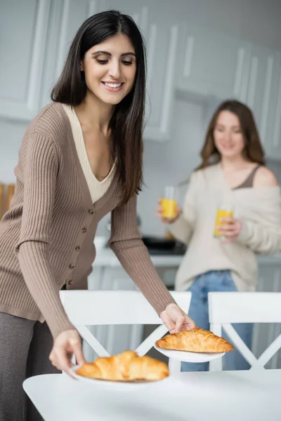 Positive lesbian woman putting croissants on table near blurred girlfriend in kitchen — Stock Photo
