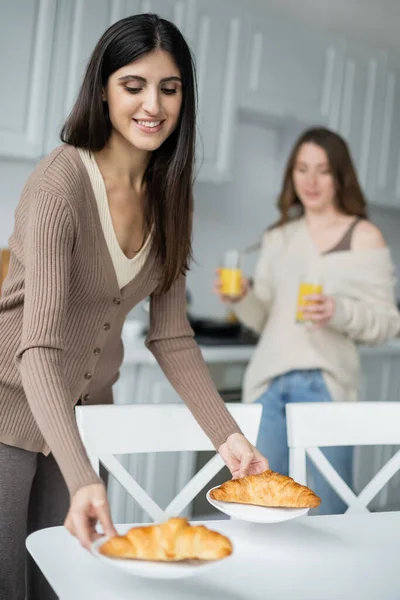 Happy woman putting croissants on table near blurred partner in kitchen — Stock Photo