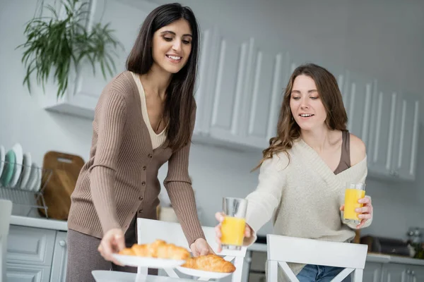Lesbianas poniendo jugo de naranja y croissants en la cocina - foto de stock