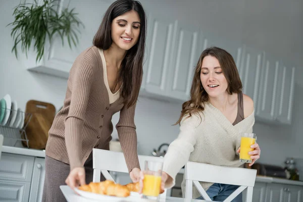 Feliz casal do mesmo sexo colocando suco de laranja e croissants na mesa na cozinha — Fotografia de Stock