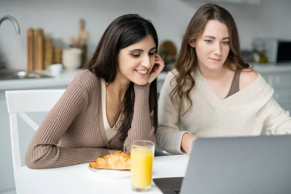 Lesbian woman looking at blurred laptop near breakfast and girlfriend in kitchen — Stock Photo