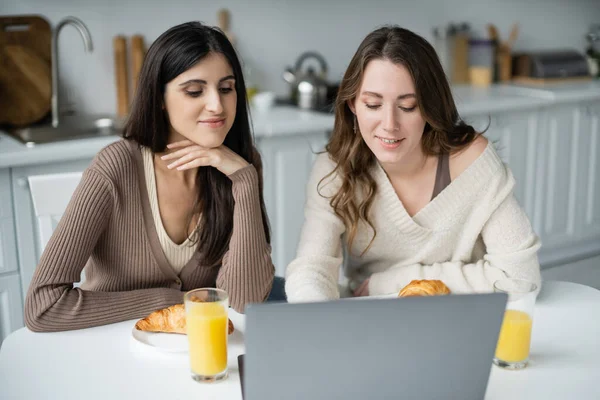Улыбающаяся лесбийская пара с ноутбуком рядом с завтраком на кухне — стоковое фото