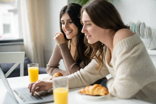 Smiling woman looking at blurred laptop near breakfast and girlfriend in sweater in kitchen — Stock Photo