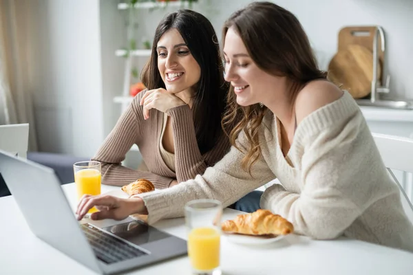 Joyeux couple de même sexe en utilisant un ordinateur portable près des croissants et du jus d'orange dans la cuisine — Photo de stock