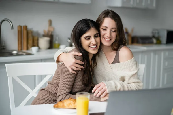 Smiling woman in sweater hugging partner near laptop and breakfast in kitchen — Stock Photo