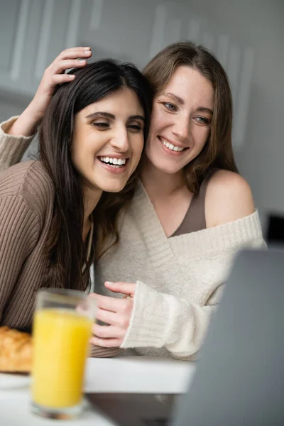 Sonriente pareja de lesbianas mirando a la computadora portátil cerca borrosa desayuno en la cocina - foto de stock