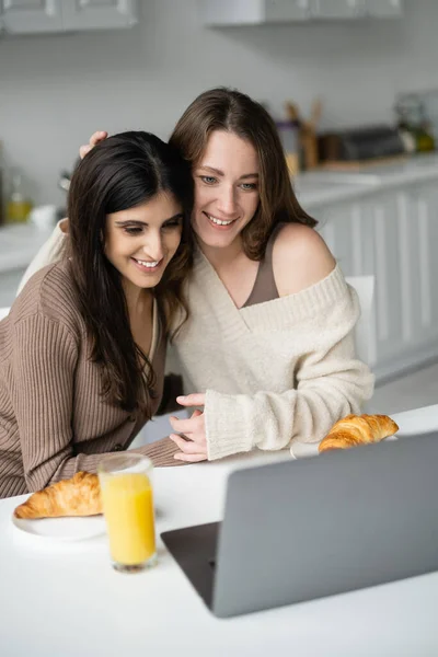 Smiling woman in sweater hugging partner near laptop and breakfast in kitchen — Stock Photo