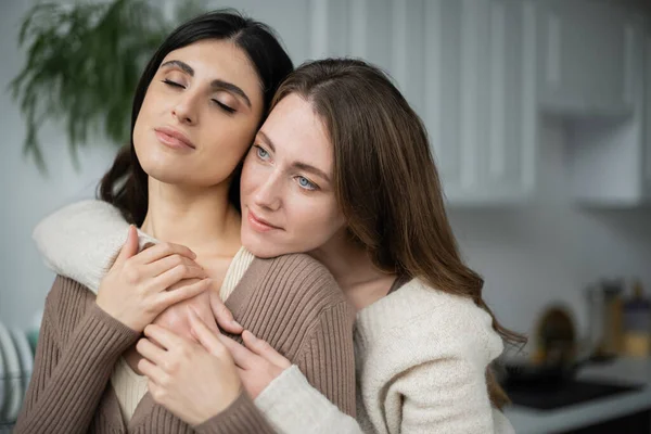 Mujer joven abrazando a su pareja lesbiana con los ojos cerrados en la cocina - foto de stock