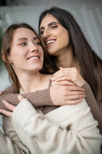 Alegre lesbiana mujer abrazando pareja en caliente suéter en casa - foto de stock