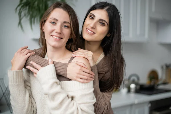 Cheerful lesbian woman hugging girlfriend in cozy sweater at home — Stock Photo