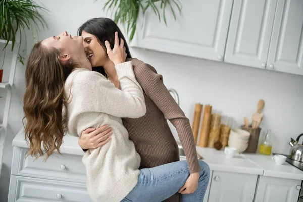Вид сбоку лесбиянки, обнимающей и целующей веселого партнера на кухне — стоковое фото