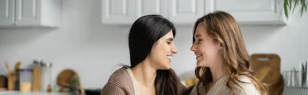 Vista lateral de pareja lesbiana positiva mirándose en la cocina, pancarta - foto de stock