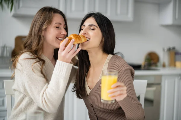 Mujer joven alimentando a su novia con croissant durante el desayuno en la cocina - foto de stock