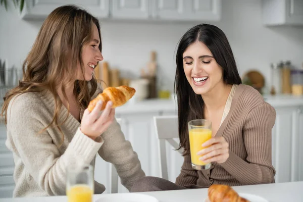 Positivo casal do mesmo sexo segurando croissant e suco de laranja durante o café da manhã na cozinha — Fotografia de Stock