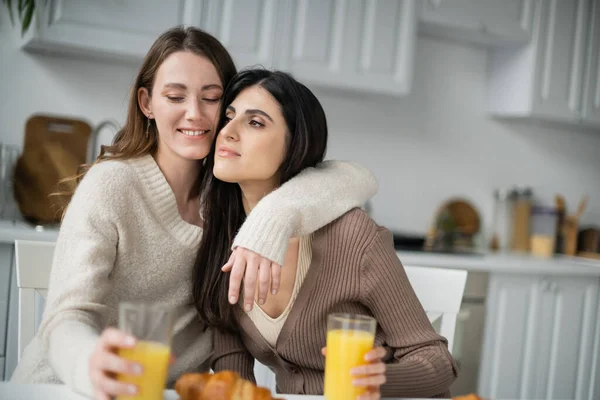 Joven lesbiana mujer abrazando novia cerca de naranja jugo y croissant en cocina - foto de stock