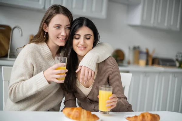 Donna positiva abbracciare fidanzata vicino croissant sfocato e succo d'arancia in cucina — Foto stock