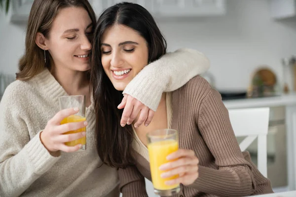 Mujer joven en suéter abrazando a su novia y sosteniendo jugo de naranja en la cocina - foto de stock