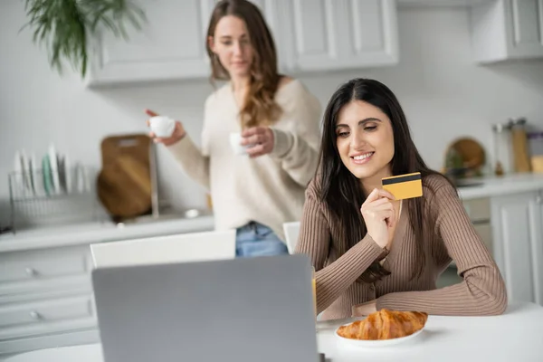 Mujer sonriente sosteniendo la tarjeta de crédito cerca del ordenador portátil y novia borrosa durante el desayuno en la cocina - foto de stock
