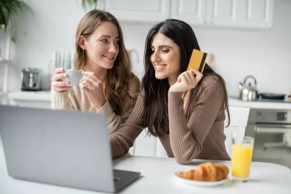 Lesbian couple using laptop and credit card during breakfast in kitchen — Stock Photo