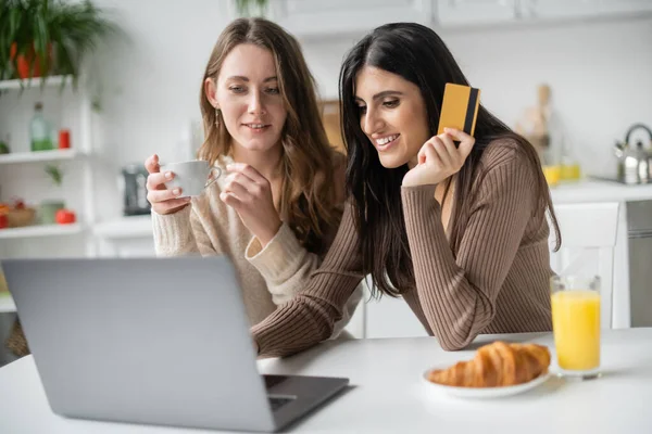 Sonriente pareja de lesbianas utilizando el ordenador portátil durante las compras en línea y el desayuno en la cocina - foto de stock