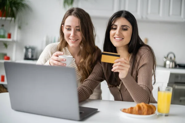 Mujer sonriente usando el ordenador portátil y la tarjeta de crédito cerca de la pareja durante el desayuno en la cocina - foto de stock