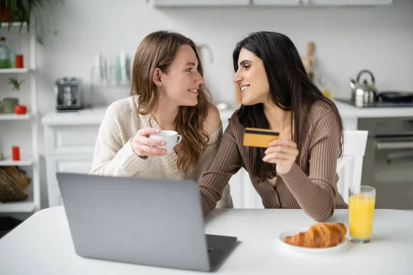 Vista lateral de sorrir casal do mesmo sexo segurando cartão de crédito e café perto de laptop e café da manhã na cozinha — Fotografia de Stock