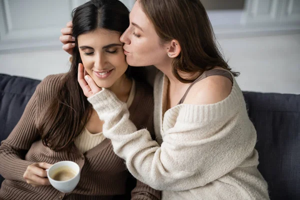 Lesbian woman in warm sweater kissing girlfriend with cup of coffee on couch — Stock Photo