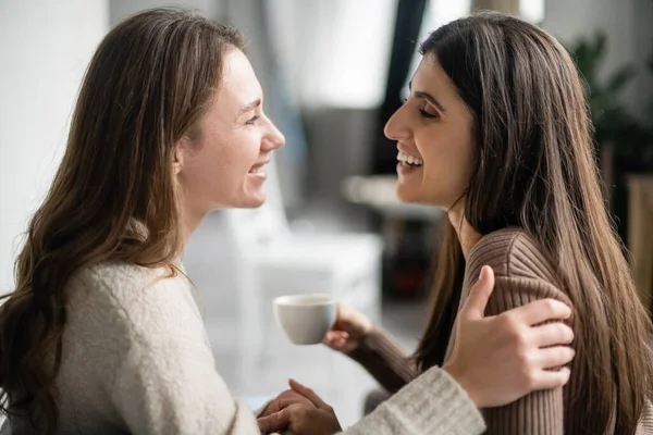 Vista lateral de mujer lesbiana en suéter abrazando y hablando con novia con taza de café en casa - foto de stock