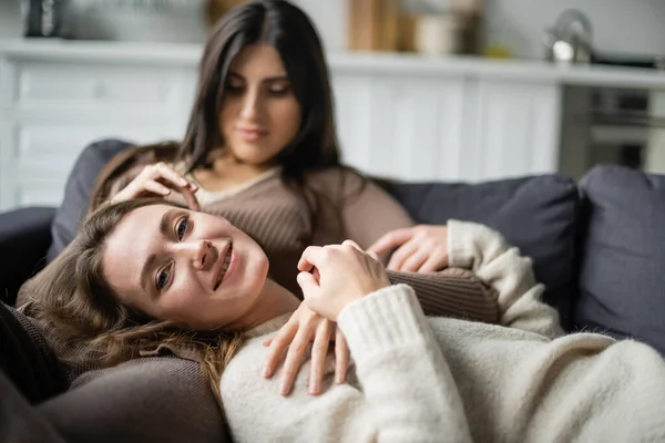 Giovane donna sorridente a macchina fotografica vicino offuscata lesbica partner sul divano — Foto stock