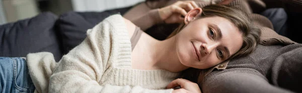 Mulher sorridente em suéter olhando para a câmera enquanto deitado perto da namorada no sofá, banner — Fotografia de Stock