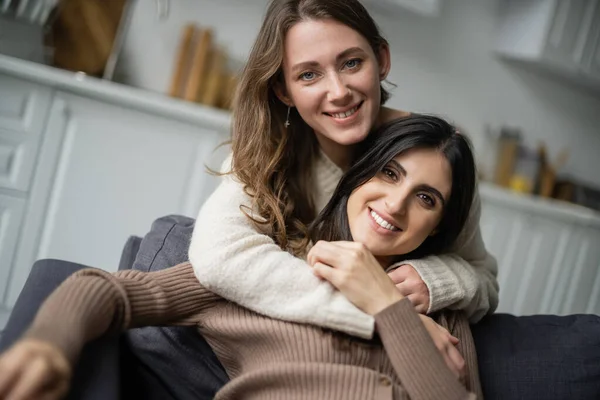 Mujer sonriente abrazando a su pareja lesbiana y mirando a la cámara en el sofá - foto de stock